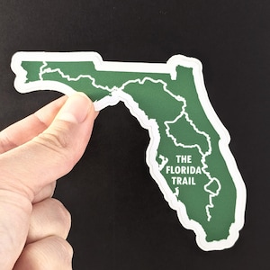 May include: A green sticker in the shape of Florida, with a white border. The text "THE FLORIDA TRAIL" is printed in white. The sticker is held by a hand against a black background.