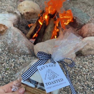 May include: A white bag with a black and white checkered ribbon tied around it. The bag contains a s'more with a graham cracker, chocolate, and marshmallow. A white tag with black text reads "Let's Get Toasted Cozy's Bachelorette".