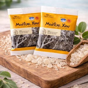 Może przedstawiać: Dwa żółto-białe opakowania produktu "Mastixa Chiou", każde po 10g, leżą na drewnianej desce. Opakowania mają tekst w języku greckim i arabskim. Małe, przezroczyste kawałki żywicy są rozrzucone.