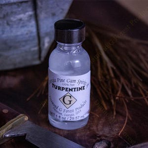 Puede incluir: Una botella de vidrio transparente con una tapa negra que contiene 29,57 ml de aguarrás puro de 100% de espíritu de goma de pino. La etiqueta dice "Diamond G Forest Products" y "Pine Gum Spirits of Turpentine".