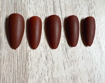 May include: A set of five almond-shaped press-on nails in a solid matte dark brown finish. The nails are arranged in a horizontal row, displaying a smooth, opaque texture and a consistent deep brown color across each individual nail piece.