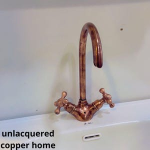 Può includere: Rubinetto da cucina in rame non laccato con due maniglie. Il rubinetto ha un becco ad arco alto ed è montato su un lavello in porcellana bianca.