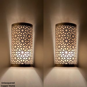 Peut inclure: Deux appliques murales cylindriques avec un motif géométrique en étoile. Les appliques sont en métal foncé et émettent une lumière chaude. Le texte "Unlacquered Copper Home" est visible en bas de l'image.