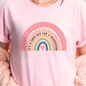 A woman wearing a pink shirt with a boho rainbow in muted colors. Between two of the rainbow stripes, it says It's a good day for a revolution.
