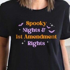 May include: Black t-shirt featuring the text "Spooky Nights & 1st Amendment Rights" in orange and purple, with bat and star graphics. A Halloween-themed graphic tee.
