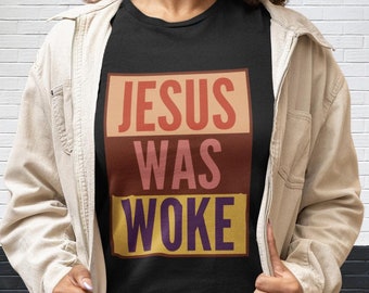 May include: A person wearing a black t-shirt with the text "JESUS WAS WOKE" in a colorful block design. The person is wearing a beige jacket and smiling.