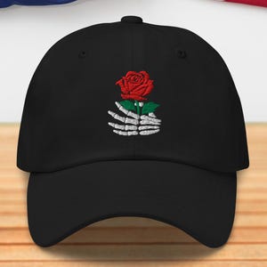 Könnte beinhalten: Schwarze Baseballkappe mit einem gestickten Design einer roten Rose, die von einer Skeletthand gehalten wird. Die Kappe befindet sich vor dem Hintergrund der amerikanischen Flagge und einer Holzoberfläche.
