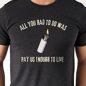 May include: Dark gray t-shirt with the text "ALL YOU HAD TO DO WAS" above a white lighter with a flame, and "PAY US ENOUGH TO LIVE" below. The shirt is a crew neck and short-sleeved.