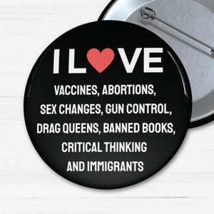 Può includere: Un bottone bianco e nero con il testo "I LOVE" in bianco con un cuore rosso al centro. Il testo sotto il cuore dice "VACCINES, ABORTIONS, SEX CHANGES, GUN CONTROL, DRAG QUEENS, BANNED BOOKS, CRITICAL THINKING AND IMMIGRANTS".