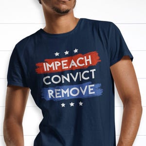 Puede incluir: Una camiseta azul marino de manga corta con un diseño gráfico que incluye el texto IMPEACH CONVICT REMOVE en blanco y rojo. El texto se superpone a trazos de pincel rojos y azules, acompañado de seis pequeñas estrellas blancas arriba y abajo.