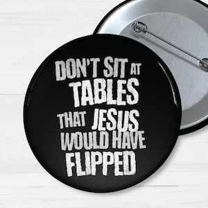 May include: A black button with the text "DON'T SIT AT TABLES THAT JESUS WOULD HAVE FLIPPED" in white, distressed font. A second button with a silver pin back is also visible.