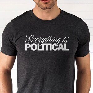 Man wearing a dark gray heather t-shirt with the phrase “Everything is Political” printed in white lettering across the chest. The word “Everything is” is written in a cursive script font, and “POLITICAL” appears in bold block capital letters underneath. The shirt is shown from shoulders to hips against a light neutral background.