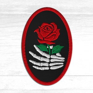Puede incluir: Parche bordado con una rosa roja y un ojo, sostenida por una mano esquelética blanca. El diseño está sobre un fondo ovalado negro con un borde rojo. Ideal para ropa o accesorios.