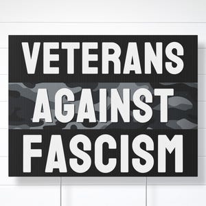 May include: A black rectangular sign with white text that reads "VETERANS AGAINST FASCISM." The text is in a bold, sans-serif font. The words are separated by a camouflage pattern. The background is a white, horizontal-slat wall.
