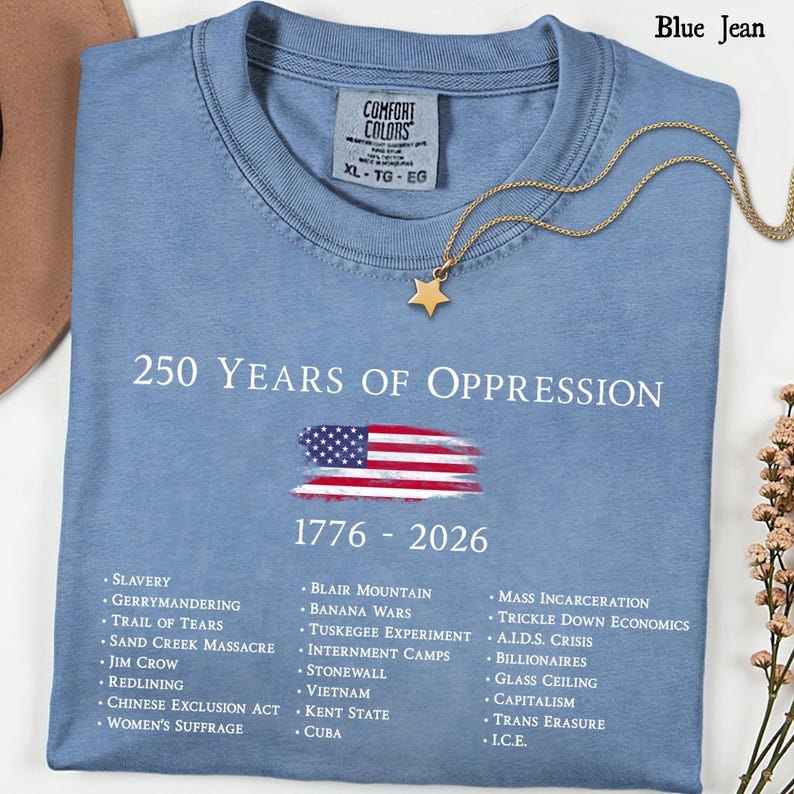 Pode incluir: Uma t-shirt azul jeans com o texto branco 250 YEARS OF OPPRESSION 1776 - 2026, um gr&aacute;fico da bandeira americana e uma lista de eventos hist&oacute;ricos incluindo Slavery, Gerrymandering, Trail of Tears e Mass Incarceration.