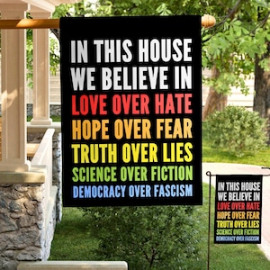 May include: A black garden flag with rainbow text that reads "In this house we believe in love over hate, hope over fear, truth over lies, science over fiction, democracy over fascism."