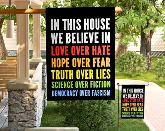 May include: A black garden flag with rainbow text that reads "In this house we believe in love over hate, hope over fear, truth over lies, science over fiction, democracy over fascism."