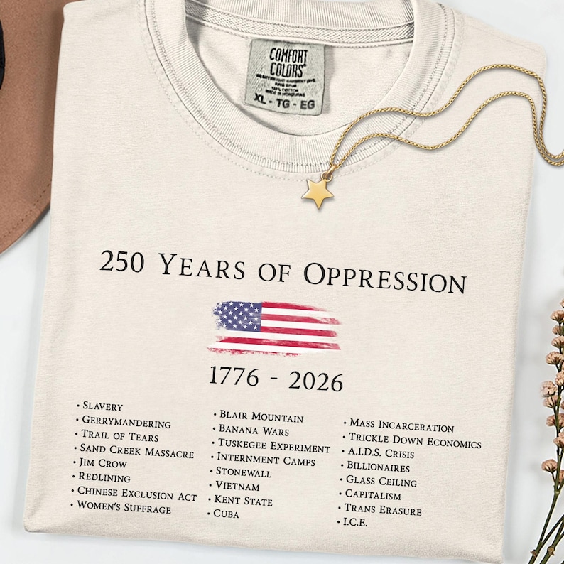 Ivory Comfort Colors t-shirt featuring “250 Years of Oppression 1776–2026” with a distressed American flag and a list of historical injustices, styled as a statement piece for America’s 250 year anniversary