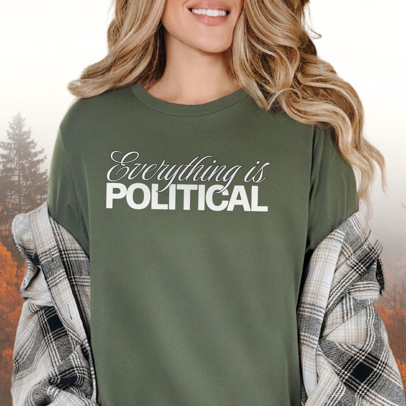 May include: Olive green t-shirt with the text "Everything is POLITICAL" in white script and block letters. The shirt is worn with a black and white plaid shirt. The background is a blurred autumn scene.
