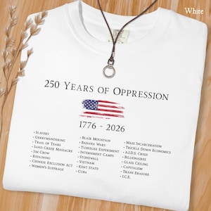 Pode incluir: Uma t-shirt branca com o texto 250 YEARS OF OPPRESSION 1776 - 2026, um gr&aacute;fico da bandeira americana desgastada e uma lista de eventos hist&oacute;ricos como escravid&atilde;o, Jim Crow e a crise da SIDA, com um colar de cord&atilde;o castanho escuro por cima.