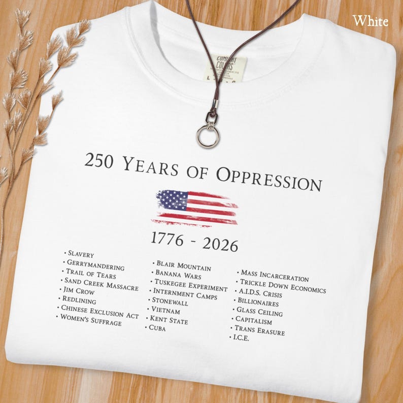 Pode incluir: Uma t-shirt branca com o texto 250 YEARS OF OPPRESSION 1776 - 2026, um gr&aacute;fico da bandeira americana desgastada e uma lista de eventos hist&oacute;ricos como escravid&atilde;o, Jim Crow e a crise da SIDA, com um colar de cord&atilde;o castanho escuro por cima.