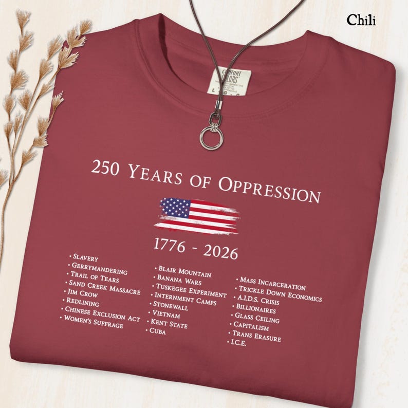 Pode incluir: Uma t-shirt vermelho-chili com o texto branco 250 YEARS OF OPPRESSION 1776 - 2026, um gr&aacute;fico de uma bandeira americana desgastada e uma lista de eventos hist&oacute;ricos incluindo Slavery, Jim Crow, Redlining e Mass Incarceration em fonte branca.