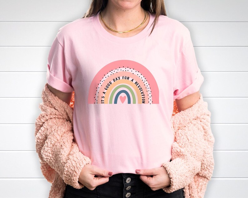 A woman wearing a pink shirt with a boho rainbow in muted colors. Between two of the rainbow stripes, it says It's a good day for a revolution.