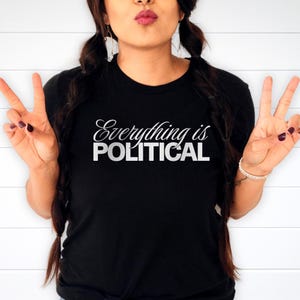 May include: Black crew neck t-shirt with the white text "Everything is POLITICAL". The shirt is made of a soft material. The person is making a peace sign with their fingers.