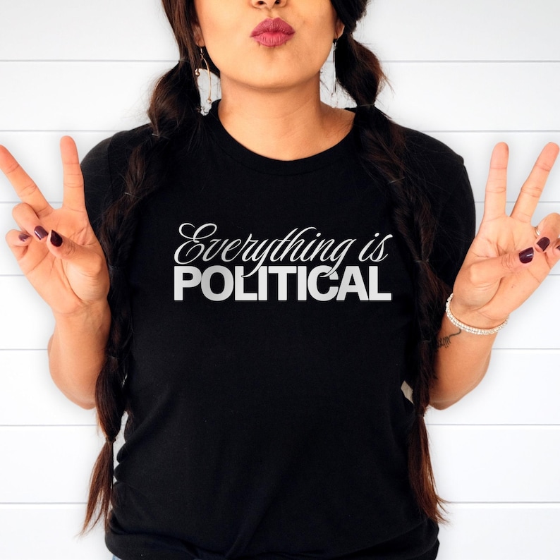May include: Black crew neck t-shirt with the white text "Everything is POLITICAL". The shirt is made of a soft material. The person is making a peace sign with their fingers.
