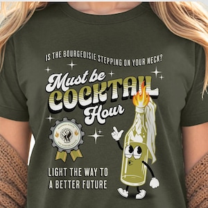 May include: Green t-shirt featuring a retro-style graphic of a Molotov cocktail with a lit fuse. The text reads "Must be Cocktail Hour" and "Light the way to a better future".