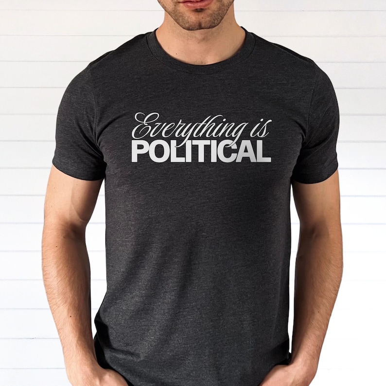 Man wearing a dark gray heather t-shirt with the phrase “Everything is Political” printed in white lettering across the chest. The word “Everything is” is written in a cursive script font, and “POLITICAL” appears in bold block capital letters underneath. The shirt is shown from shoulders to hips against a light neutral background.