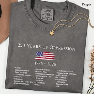 Pode incluir: Uma t-shirt cinza-carv&atilde;o com o texto branco 250 YEARS OF OPPRESSION, uma bandeira americana desgastada, os anos 1776 - 2026 e uma lista de eventos hist&oacute;ricos incluindo Slavery, Jim Crow e Mass Incarceration impressos em branco.