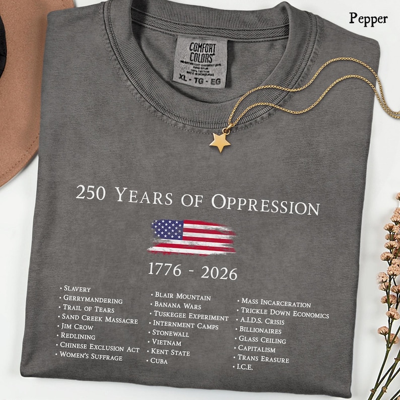Pode incluir: Uma t-shirt cinza-carv&atilde;o com o texto branco 250 YEARS OF OPPRESSION, uma bandeira americana desgastada, os anos 1776 - 2026 e uma lista de eventos hist&oacute;ricos incluindo Slavery, Jim Crow e Mass Incarceration impressos em branco.