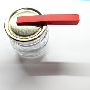 May include: A glass jar with a gold lid and a red plastic lid opener. The jar is empty and the lid opener is attached to the lid.