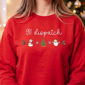 May include: A bright red crewneck sweatshirt featuring the white cursive text 911 dispatch above a row of holiday icons including holly leaves, a snowman, a blue snowflake, a green Christmas tree, a second snowflake, Santa Claus, and holly.