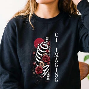 May include: A black long-sleeve sweatshirt featuring a graphic print of a human rib cage intertwined with red and pink roses. The design includes a dark red circle and the vertical white text "CT IMAGING" positioned to the right of the skeleton.