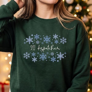 May include: A dark forest green long-sleeve sweatshirt featuring a central graphic of light blue snowflakes surrounding the white cursive text 911 dispatcher. The design is arranged in a rectangular pattern with various snowflake shapes.