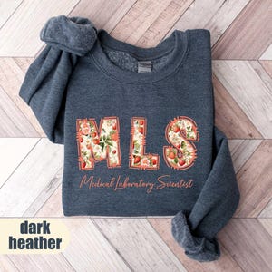 Puede incluir: Sudadera gris oscuro con las letras "MLS" en un dise&ntilde;o floral de fresas. Las palabras "Medical Laboratory Scientist" est&aacute;n impresas debajo. La sudadera est&aacute; doblada sobre una superficie de madera.