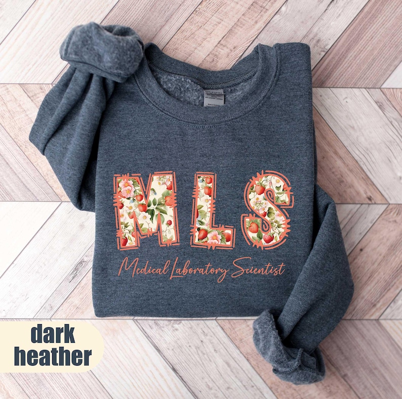 Puede incluir: Sudadera gris oscuro con las letras "MLS" en un dise&ntilde;o floral de fresas. Las palabras "Medical Laboratory Scientist" est&aacute;n impresas debajo. La sudadera est&aacute; doblada sobre una superficie de madera.