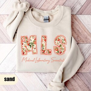 Puede incluir: Sudadera color arena con las letras "MLS" en un dise&ntilde;o floral de fresas. Las palabras "Medical Laboratory Scientist" est&aacute;n impresas debajo. La sudadera est&aacute; doblada sobre una superficie de madera.