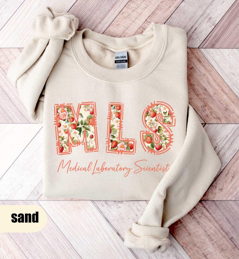 Puede incluir: Sudadera color arena con las letras "MLS" en un dise&ntilde;o floral de fresas. Las palabras "Medical Laboratory Scientist" est&aacute;n impresas debajo. La sudadera est&aacute; doblada sobre una superficie de madera.
