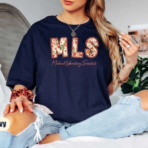 Puede incluir: Camiseta azul marino con las letras "MLS" en un dise&ntilde;o floral y las palabras "Medical Laboratory Scientist" debajo. La camiseta tiene un corte holgado.