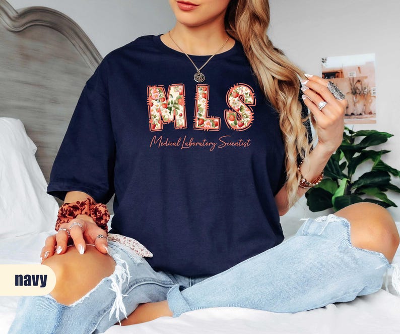 Puede incluir: Camiseta azul marino con las letras "MLS" en un dise&ntilde;o floral y las palabras "Medical Laboratory Scientist" debajo. La camiseta tiene un corte holgado.