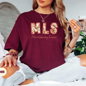 Puede incluir: Camiseta granate con el texto "MLS Medical Laboratory Scientist" en un dise&ntilde;o floral. La camiseta es de color granate liso y tiene cuello redondo.