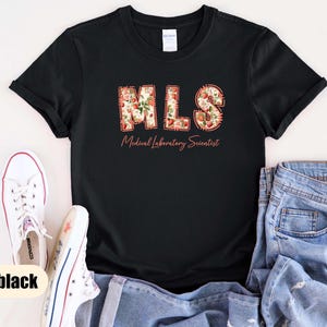 Puede incluir: Camiseta negra con las letras "MLS" en un dise&ntilde;o floral, con las palabras "Medical Laboratory Scientist" debajo. Zapatillas blancas y vaqueros azules tambi&eacute;n est&aacute;n en la imagen.