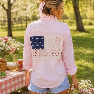 May include: A light pink long-sleeve button-down shirt featuring a ruffled collar and a large rectangular patch on the back. The patch displays a blue square with white stars and a white section decorated with small red heart-shaped embroidery.