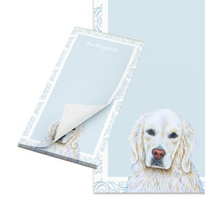 May include: Light blue notepad with a decorative border and the text "The Fitzgeralds." The notepad features a detailed illustration of a Golden Retriever dog. The top sheet is partially peeled back, revealing the adhesive.