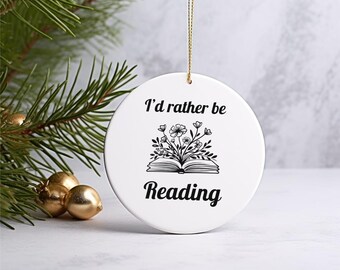 Puede incluir: Adorno redondo de cerámica blanca con el texto negro "I'd rather be Reading". El adorno presenta una ilustración negra de un libro abierto con flores y hojas. Se adjunta un hilo dorado para colgarlo.