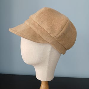 May include: A tan newsboy cap with a rounded crown and a short, stiff brim. The hat is constructed from a soft, felt-like material and features a structured band that sits above the brim, creating a layered appearance on the head.
