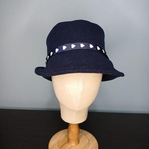 May include: A navy blue cloche hat with a white heart-patterned ribbon band. The hat is displayed on a wooden mannequin head. The hat has a slightly curved brim and a rounded crown. The ribbon band is wrapped around the base of the crown.